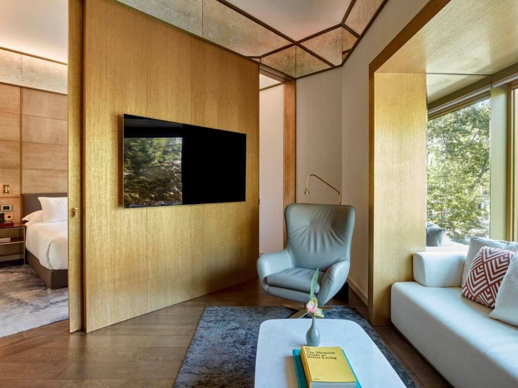 Modern Almanac Suite with a partition wall, flat-screen TV, gray armchair, white sofa, large windows, and a coffee table with books and a flower vase. Two-bedroom suite layout with bedroom visible in the background.